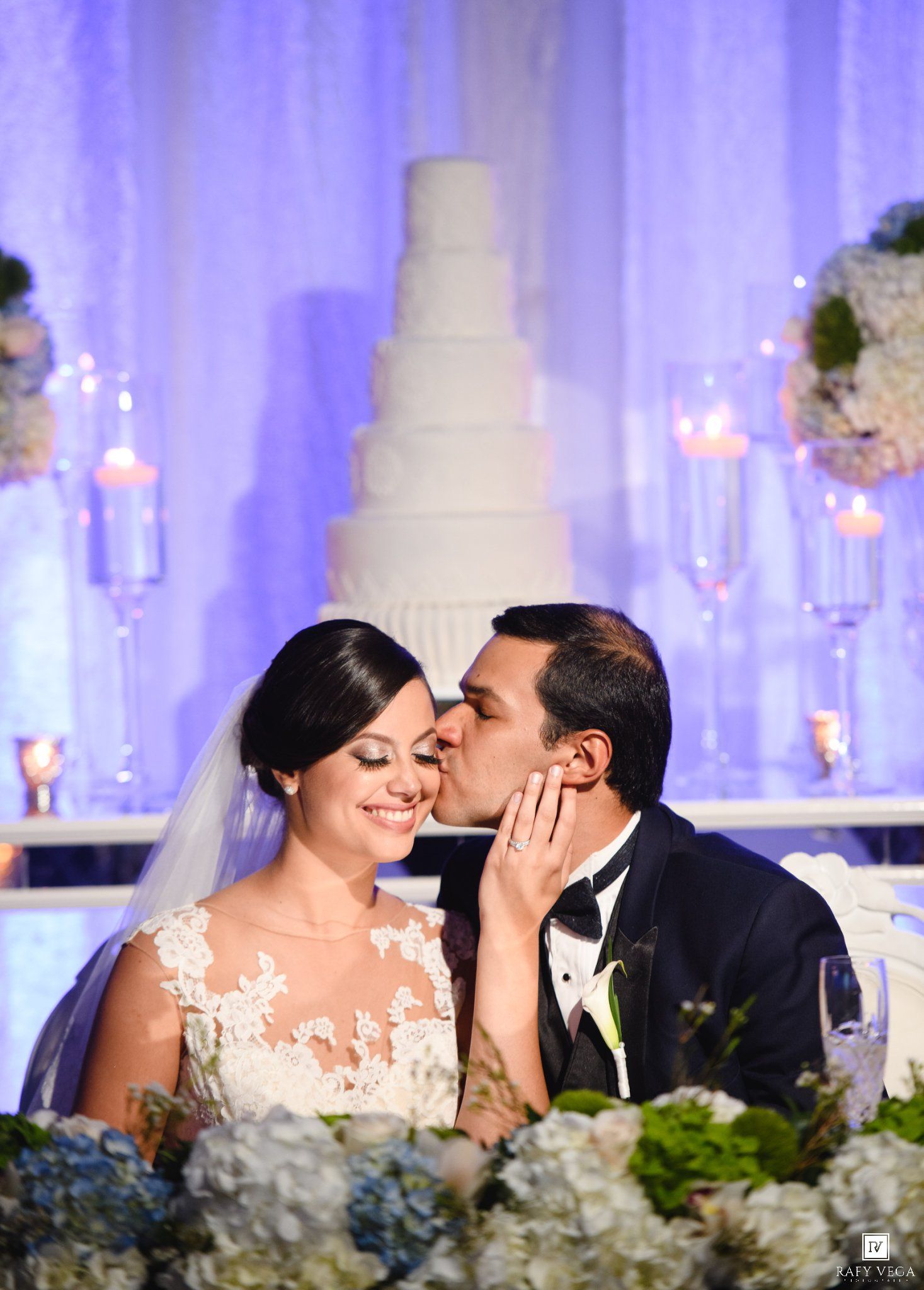 Bride and groom share a kiss at elegant wedding reception with white tiered cake and floral arrangements.