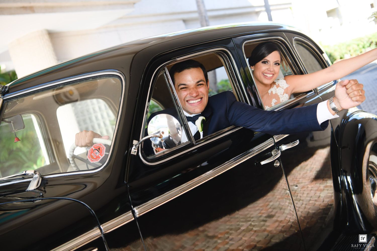Smiling bride and groom leaning out windows of a classic black wedding car on their special day.