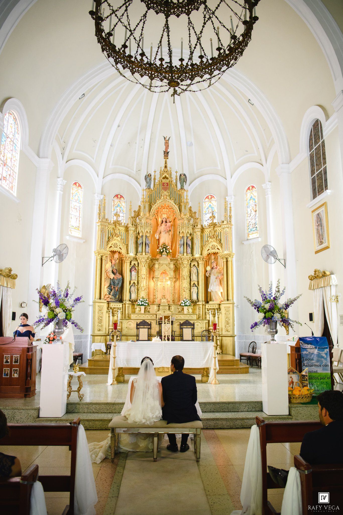 Bride and groom sit before an ornate golden altar in a white domed Catholic church during their wedding ceremony.