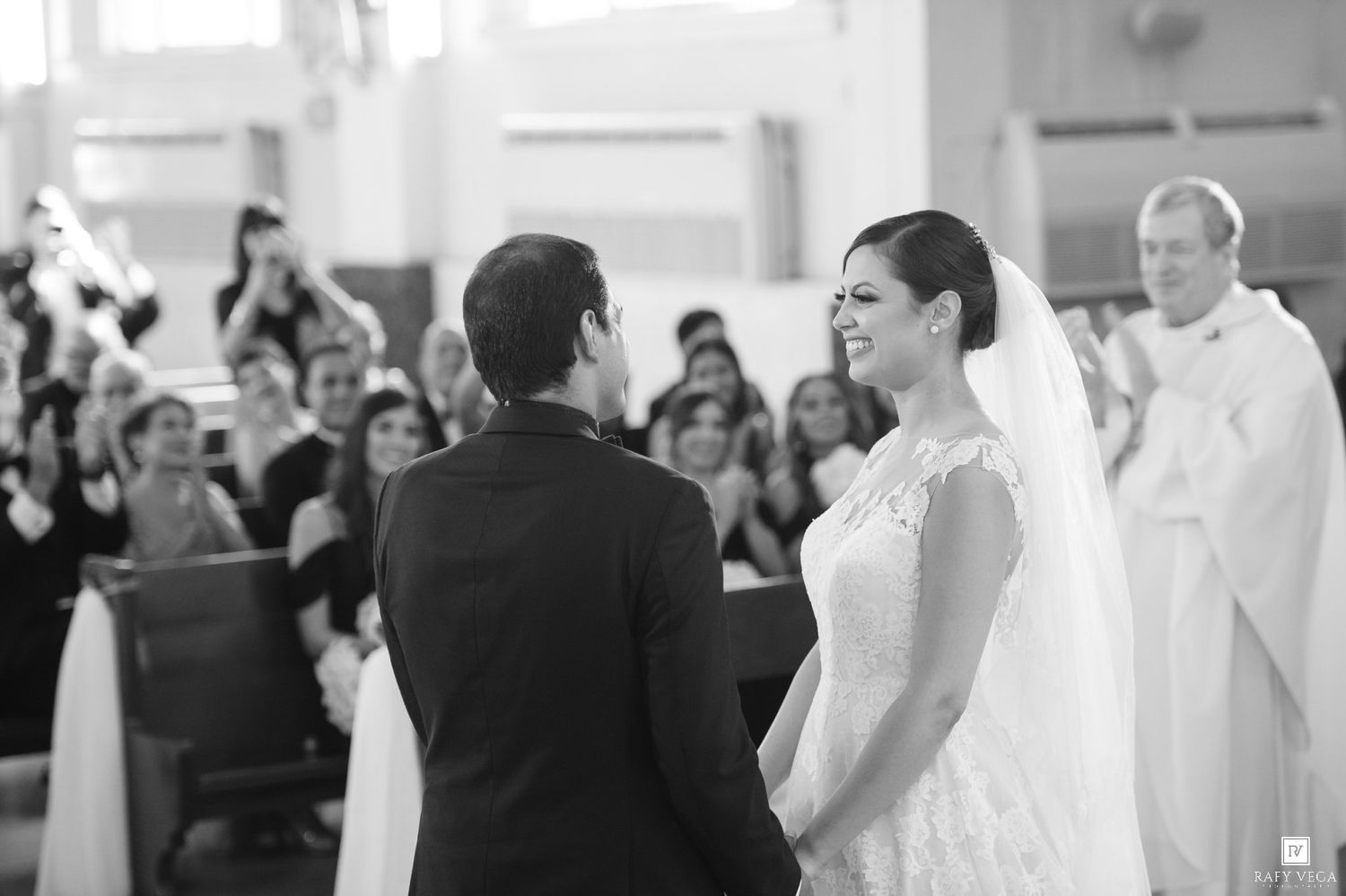Bride and groom exchange vows at church altar, smiling joyfully, with guests and priest in background.