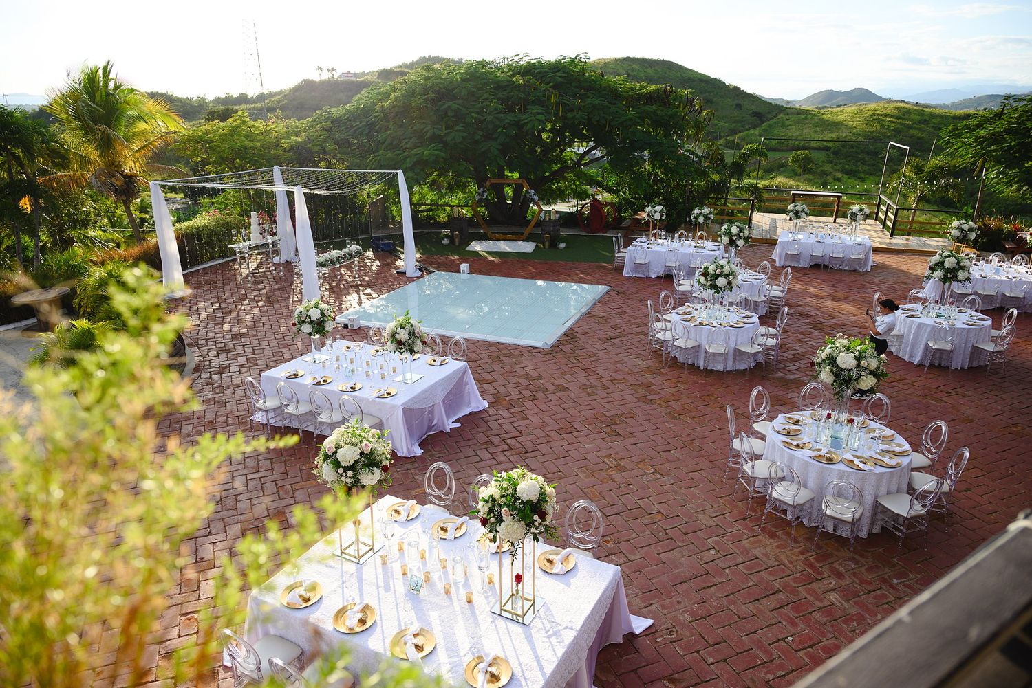 Elegant outdoor wedding reception on a brick terrace with white-draped round tables, floral centerpieces, and a pool overlooking lush hills.