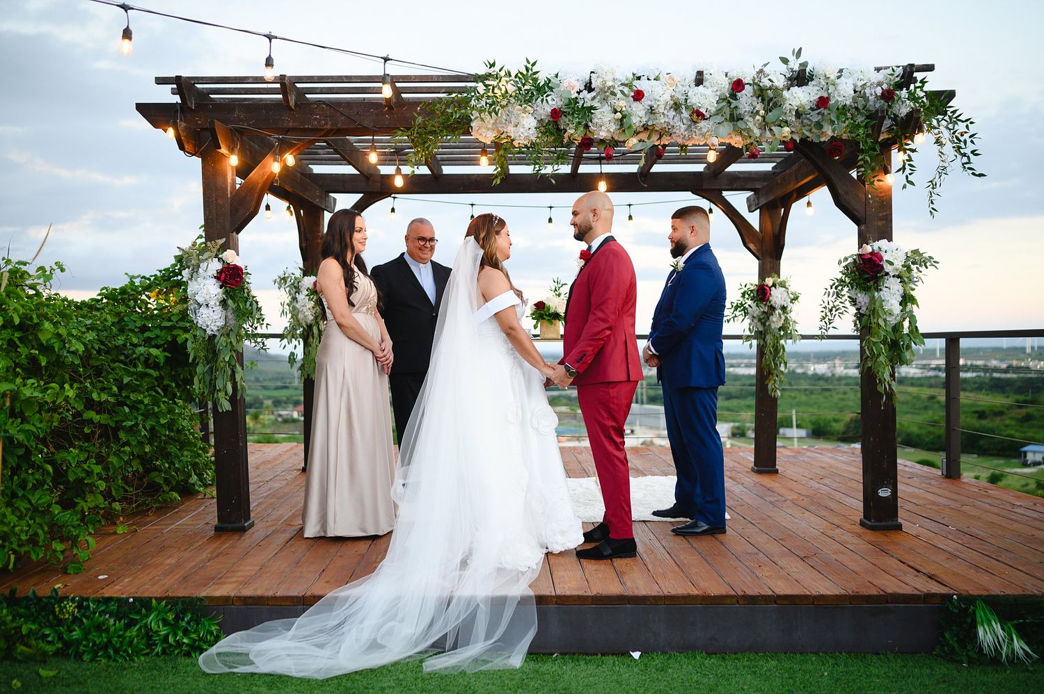 Wedding ceremony on a wooden deck pergola with floral arch, bride, groom in red suit, officiant, and wedding party.