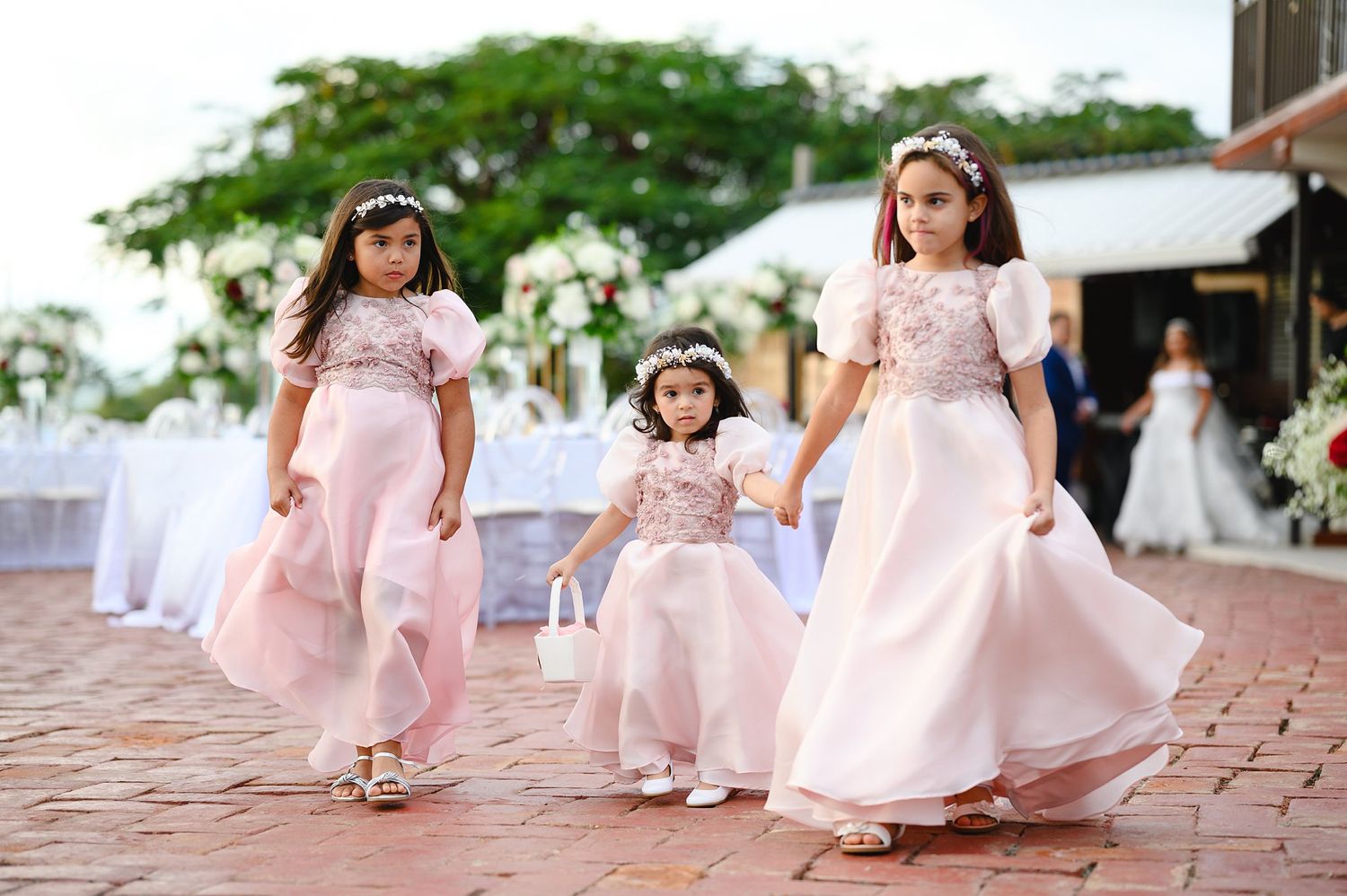 Three flower girls in pink dresses walk at an outdoor wedding ceremony, carrying a basket and wearing floral crowns.