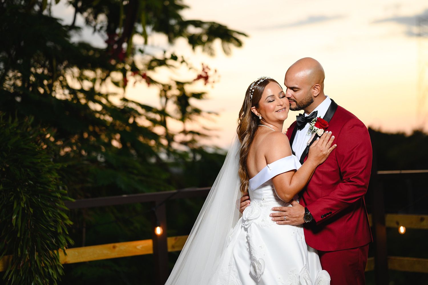 Bride and groom embrace at sunset on a wooden deck, she in a white gown, he in a red suit with bow tie.