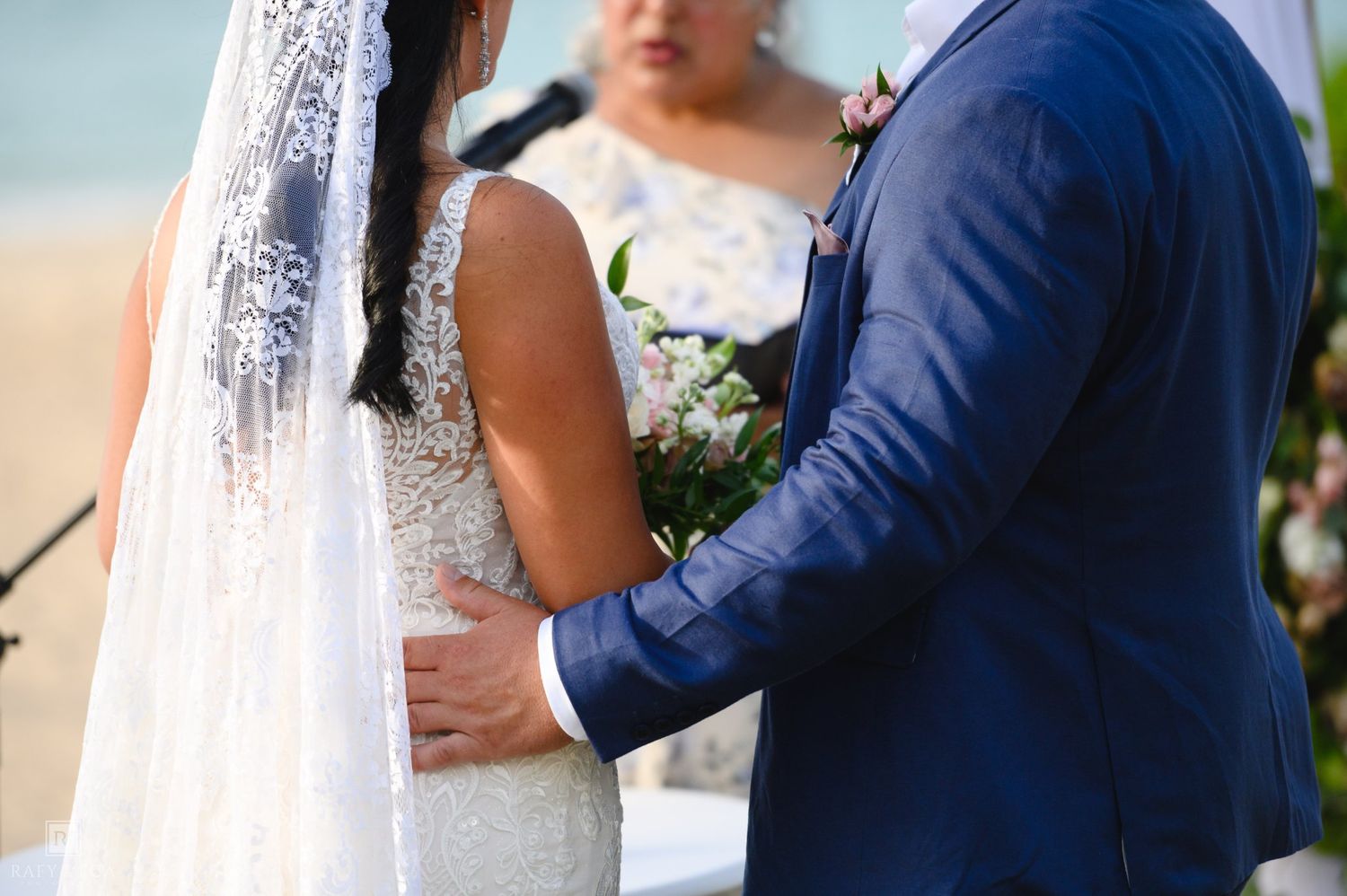 Bride and groom stand together during outdoor beach wedding ceremony, groom in navy suit rests hand on bride's waist.