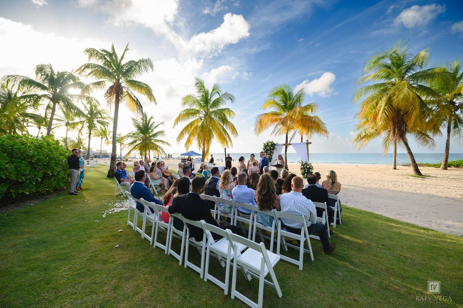 Outdoor beach wedding ceremony with guests seated on white chairs under tropical palm trees near the ocean on a sunny day.