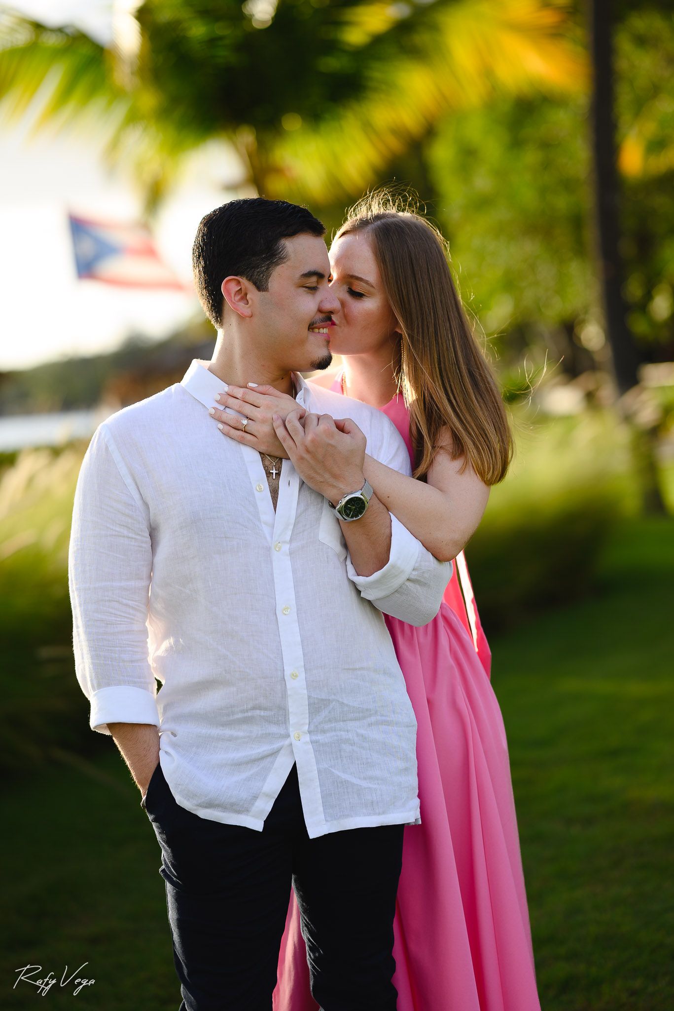 Couple sharing a romantic kiss outdoors near palm trees with a Puerto Rico flag in the background.