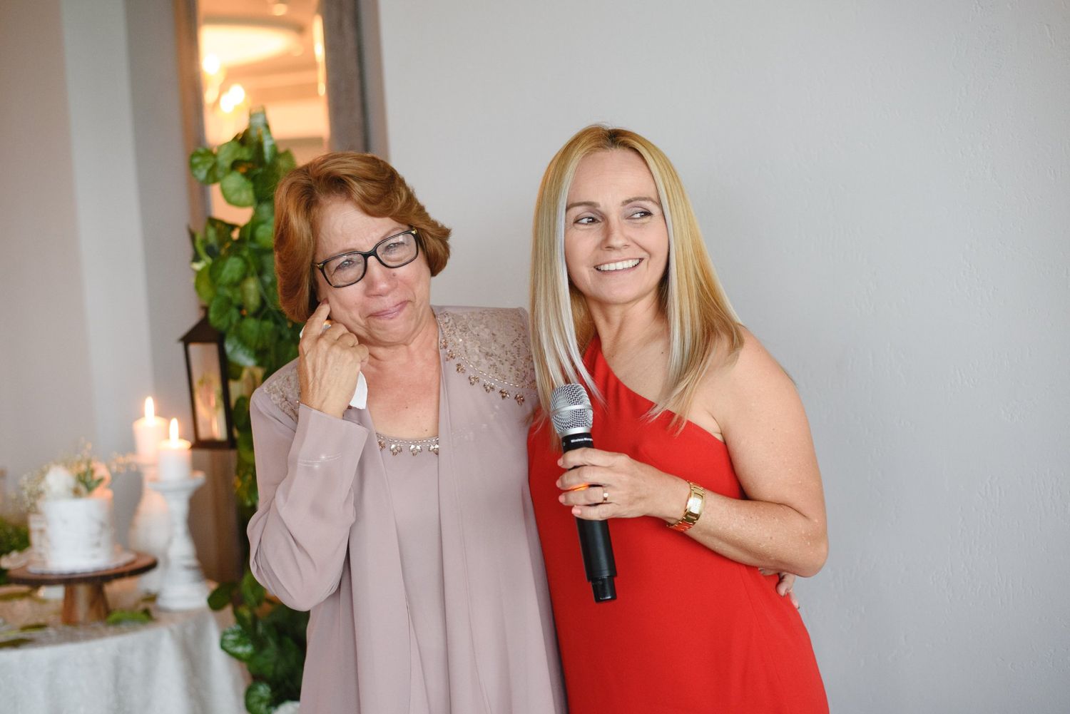 Two women posing together at wedding reception, one holding a microphone, with candles in background.
