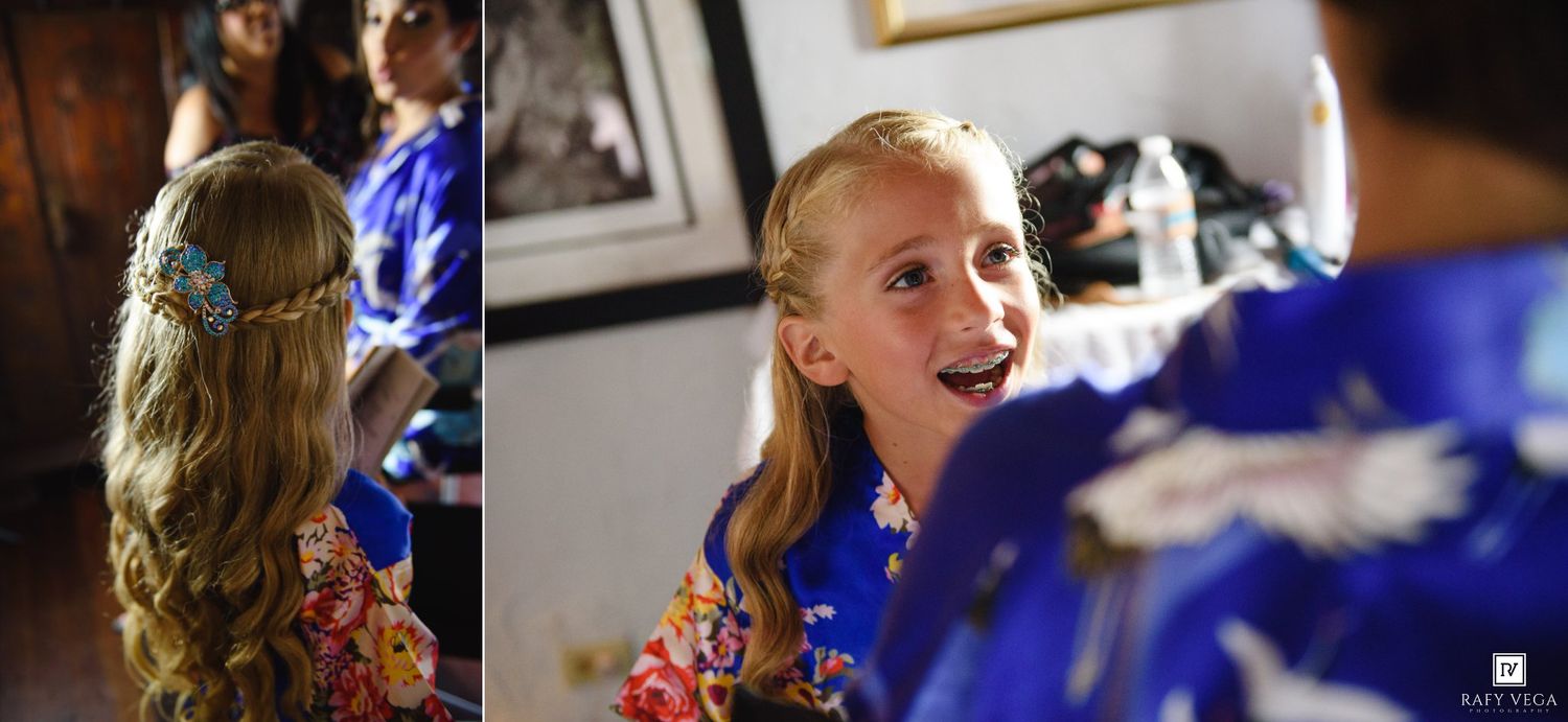 Young flower girl in blue robe with floral hair accessory laughing during wedding preparations.