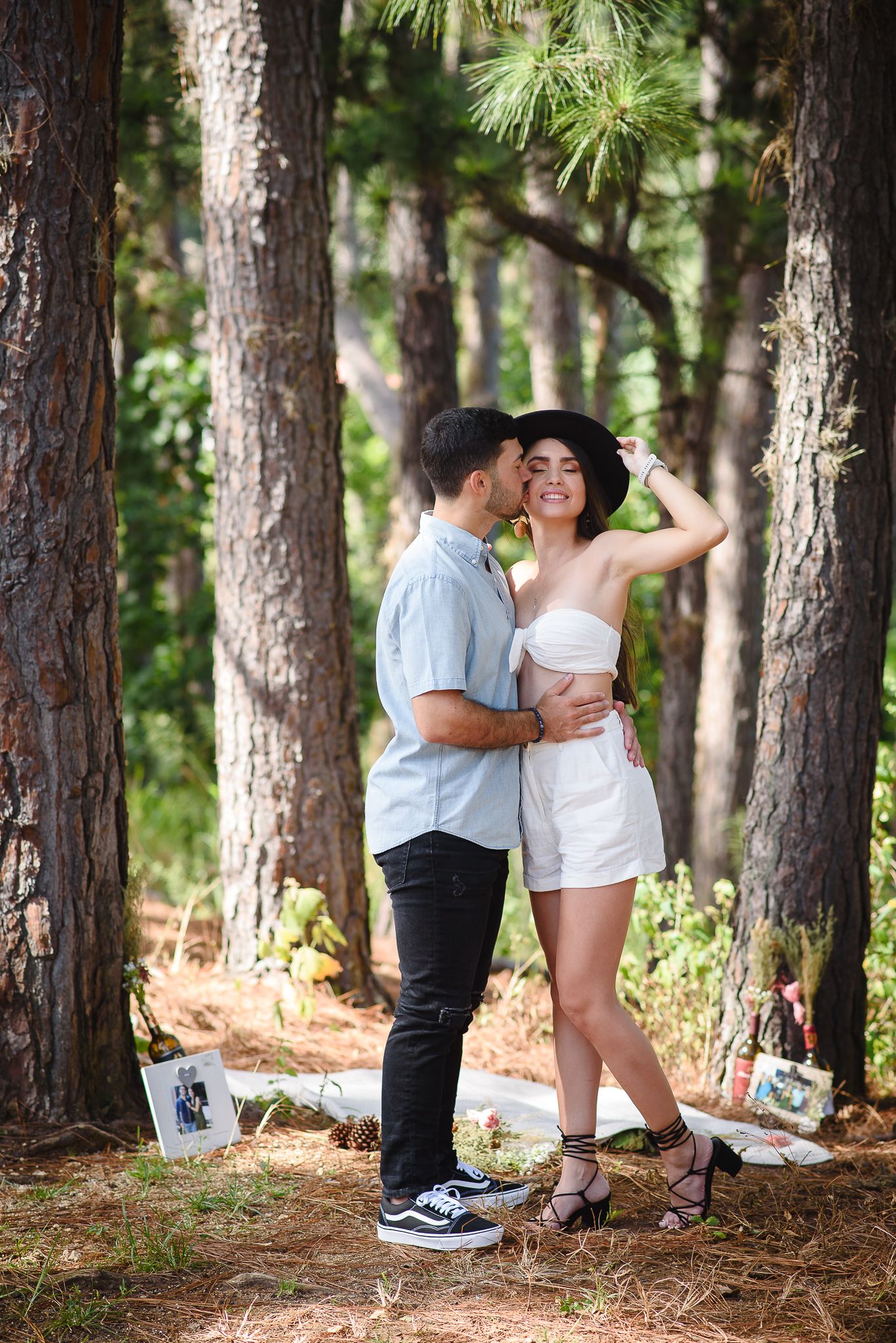 Romantic couple embracing in a pine forest, woman in white outfit and black hat, man in blue shirt and black pants.