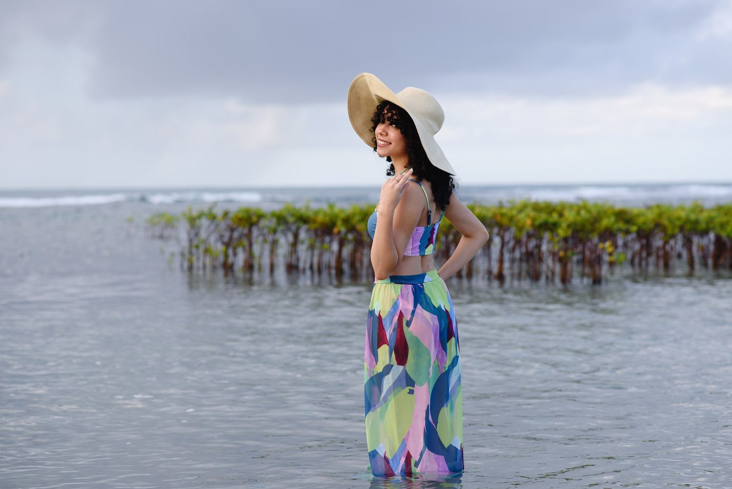 Woman in colorful maxi dress and sun hat standing in shallow water near mangrove plants on a cloudy day.