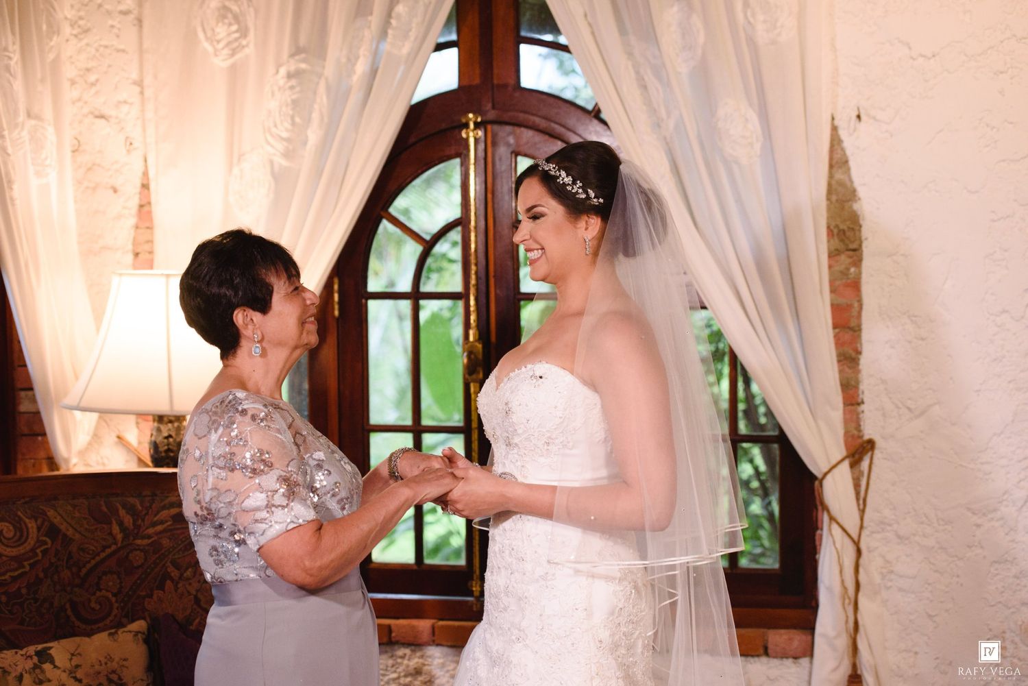 A bride in a white gown and veil holds hands with an older woman in a silver dress near a arched window.