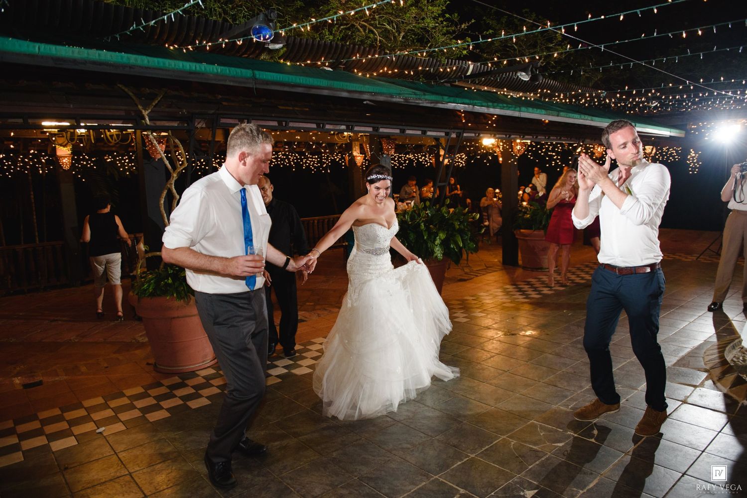 Bride and groom share first dance at outdoor wedding reception with string lights and guests celebrating.