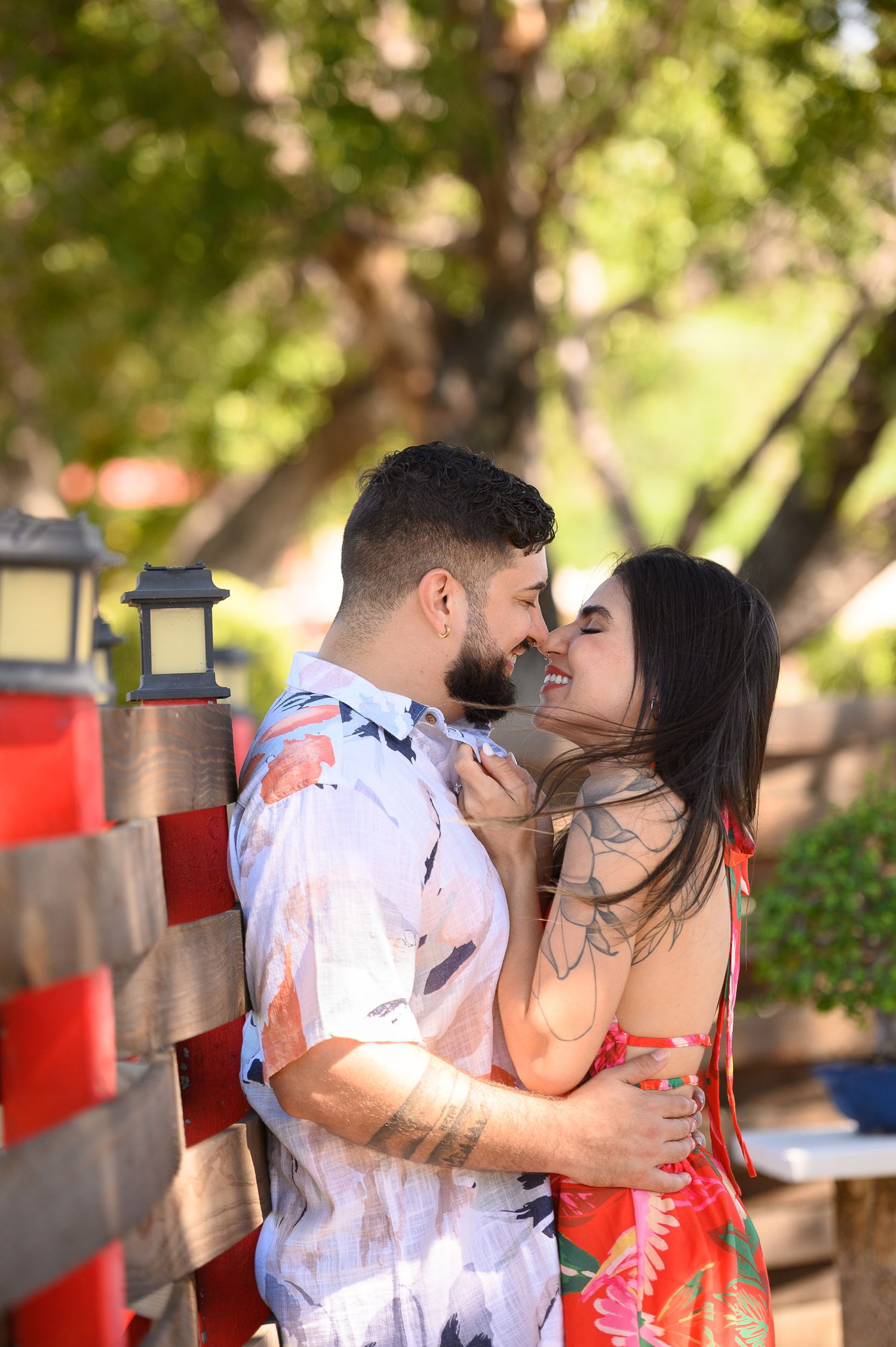 A couple shares a romantic moment, nearly kissing outdoors near a red fence with lush green trees in the background.