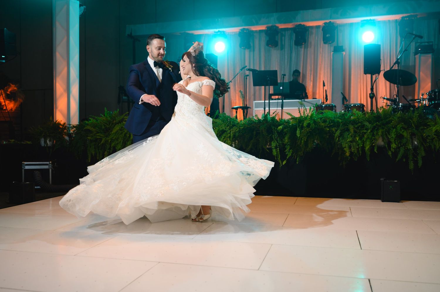 Bride and groom share romantic first dance, wedding gown flowing on white dance floor with lush greenery backdrop.