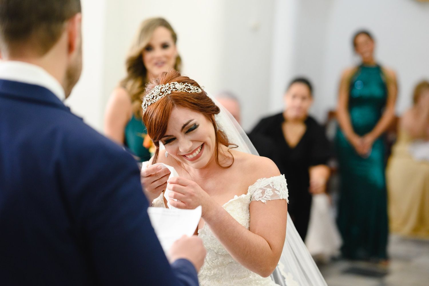 Smiling bride in white gown and tiara exchanges rings with groom during wedding ceremony, bridesmaids in green dresses behind.