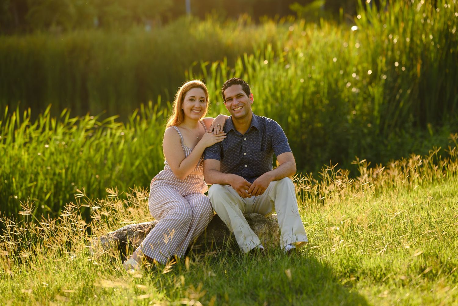 A couple sits together on a log in a sunny green meadow, smiling warmly in golden hour light.