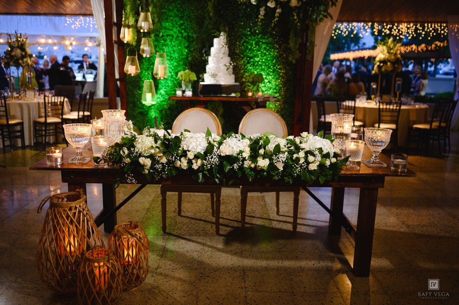 Elegant wedding sweetheart table with white floral garland, candles, and a tiered cake against a lush green moss wall backdrop.