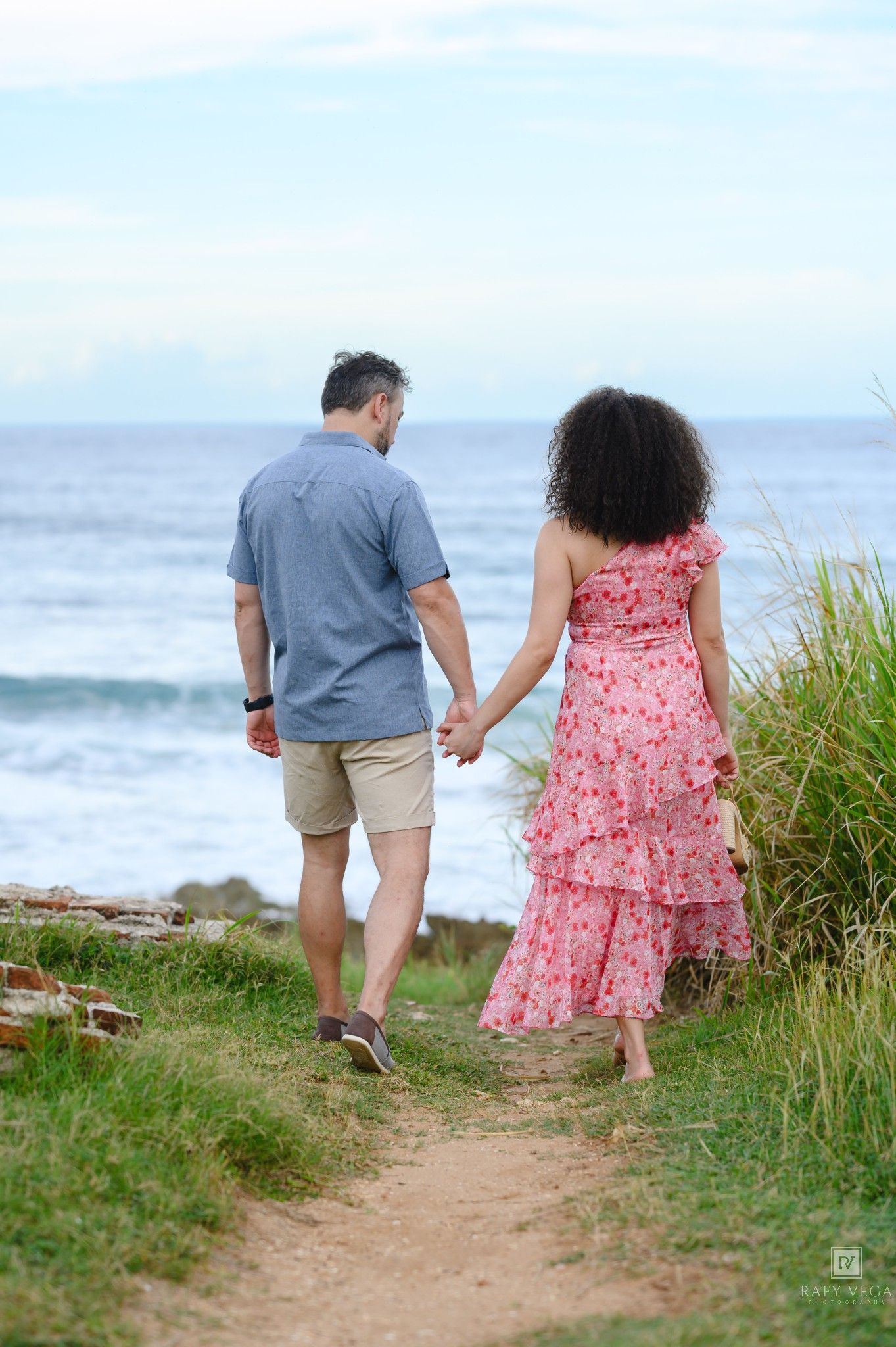 Couple holding hands walking along a coastal path toward the ocean, woman in pink floral dress, man in blue shirt.