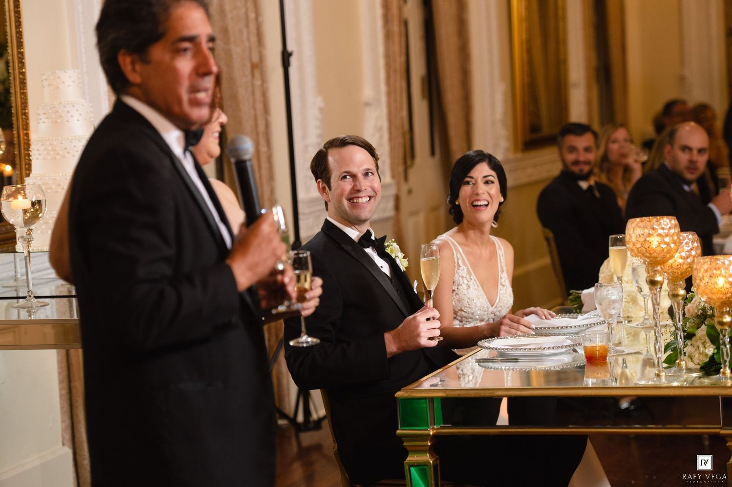 Bride and groom smile during wedding reception toast at elegant venue with candlelit table decor.
