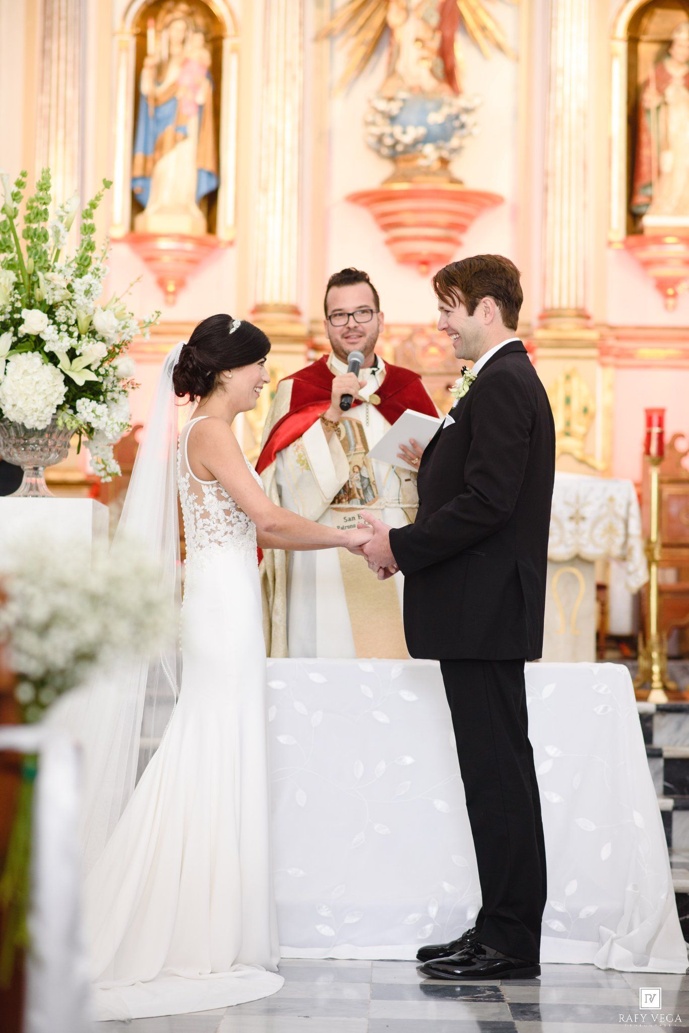Bride and groom exchange vows at a church altar, officiated by a priest in red vestments.