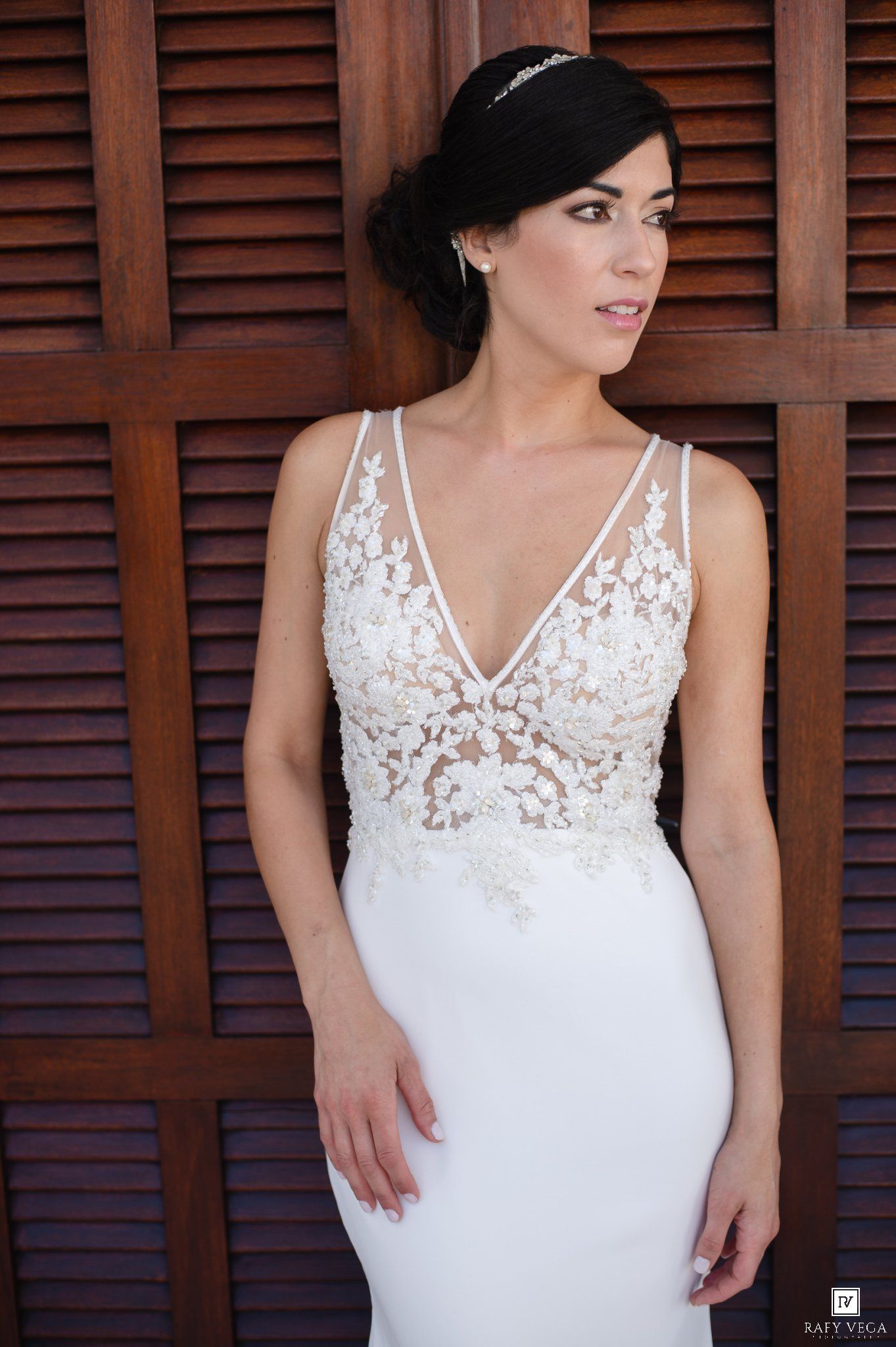 Bride in elegant lace V-neck wedding gown with floral embroidery, wearing a delicate hair accessory, posed against wooden shutters.