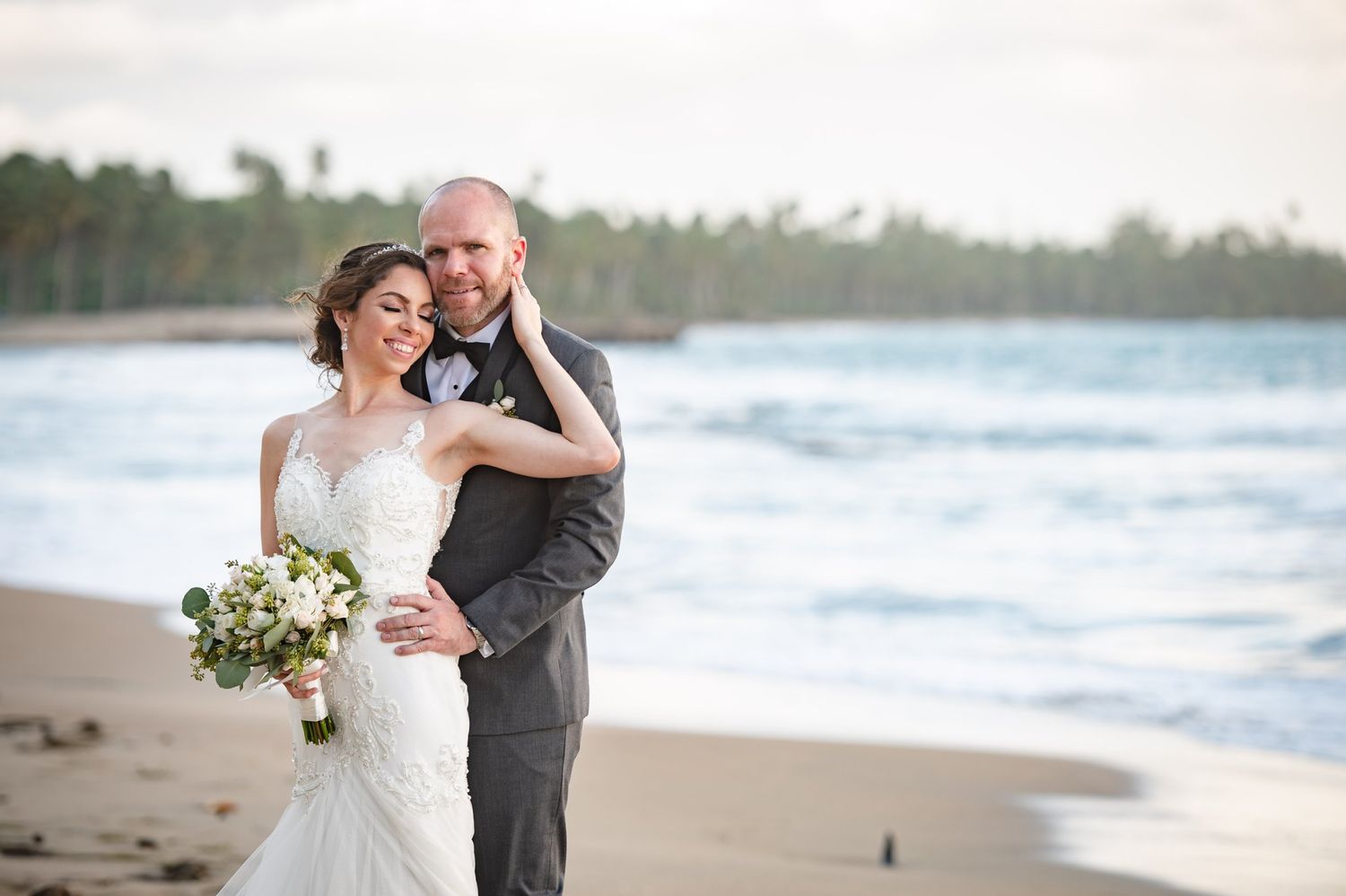 Bride and groom embrace on a tropical beach, bride holding a white bouquet in a mermaid gown, groom in gray suit.