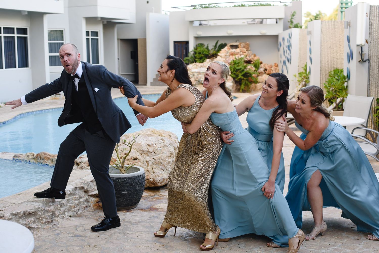 Wedding party laughing and playing near a pool, bridesmaids in blue dresses with a man in a suit.