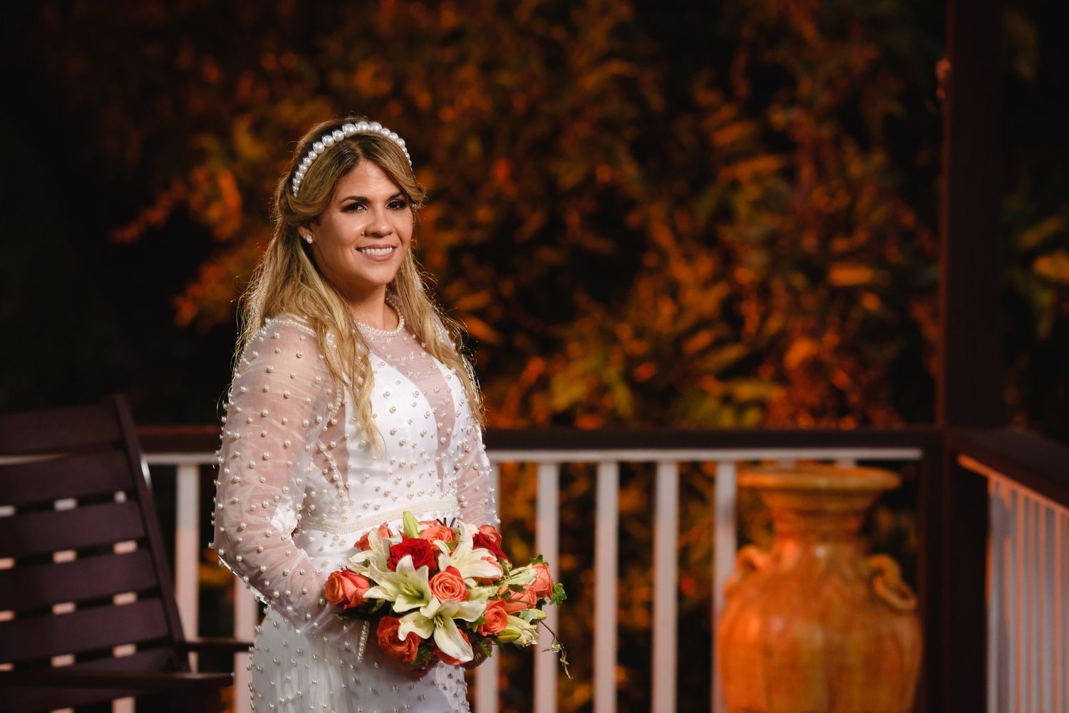 Smiling bride in lace long-sleeve wedding gown and pearl headband holding red and white floral bouquet outdoors.