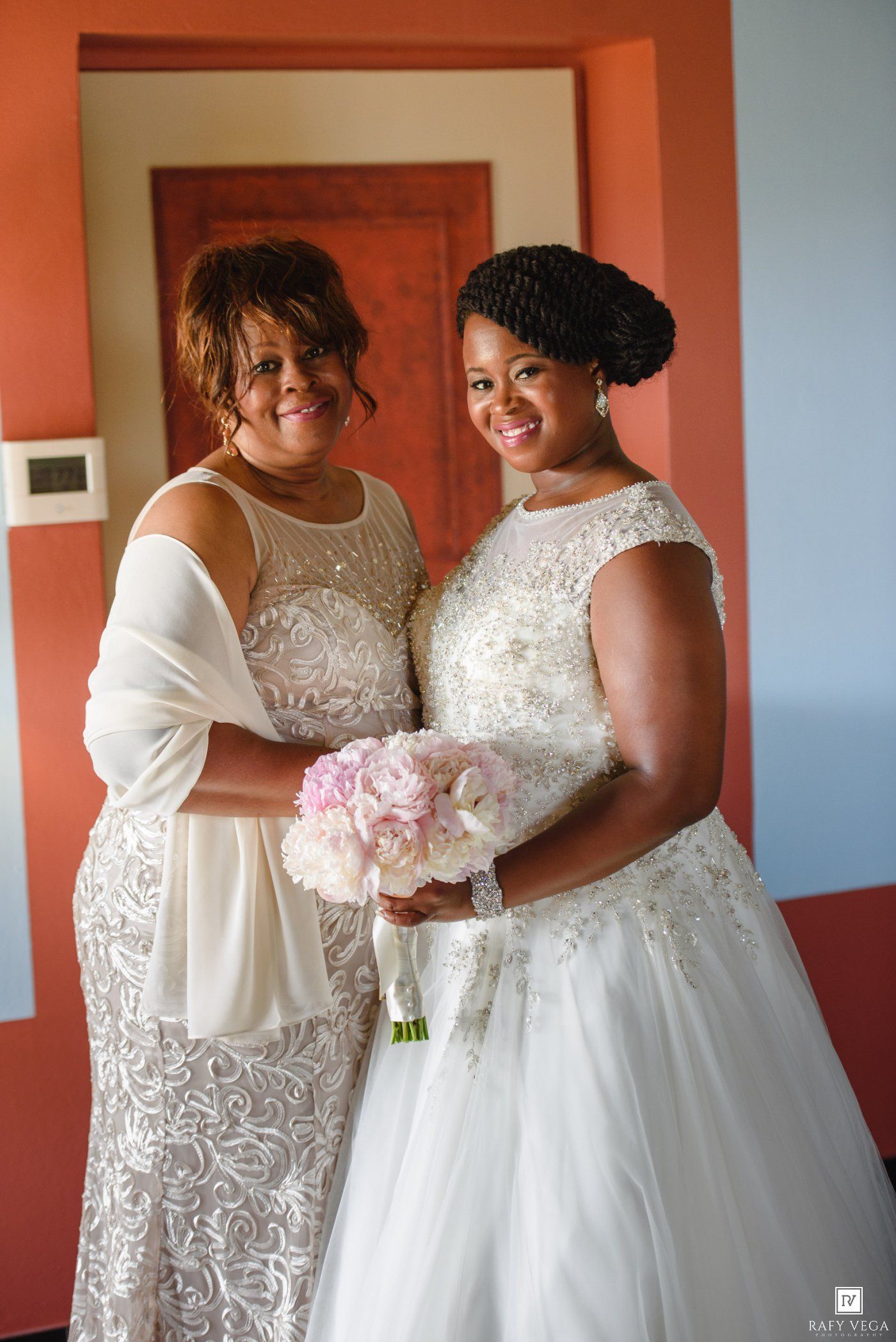 A bride in a white beaded gown holds pink flowers beside a woman in a silver dress, both smiling indoors.