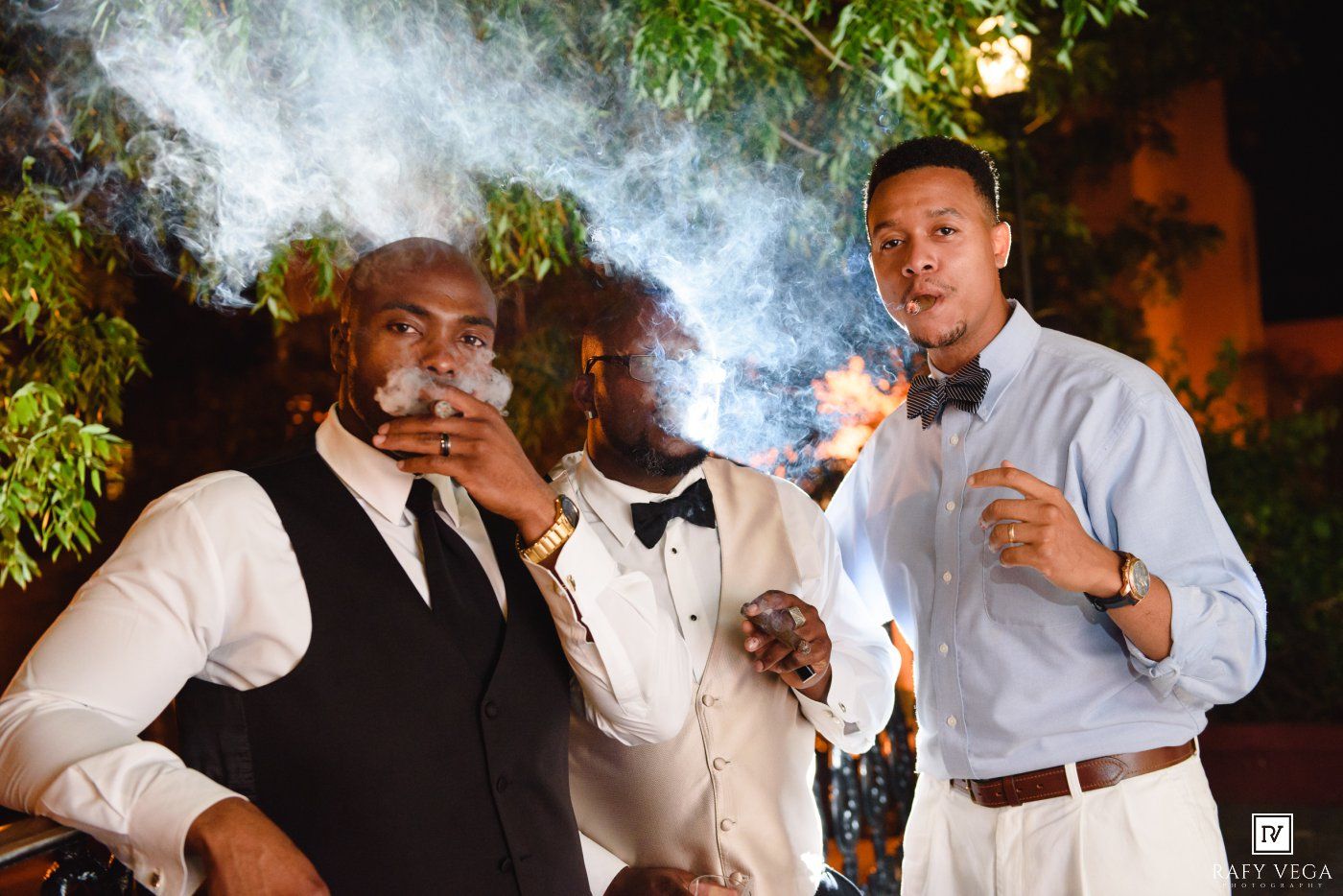 Two men in formal attire smoking cigars at an outdoor evening event.