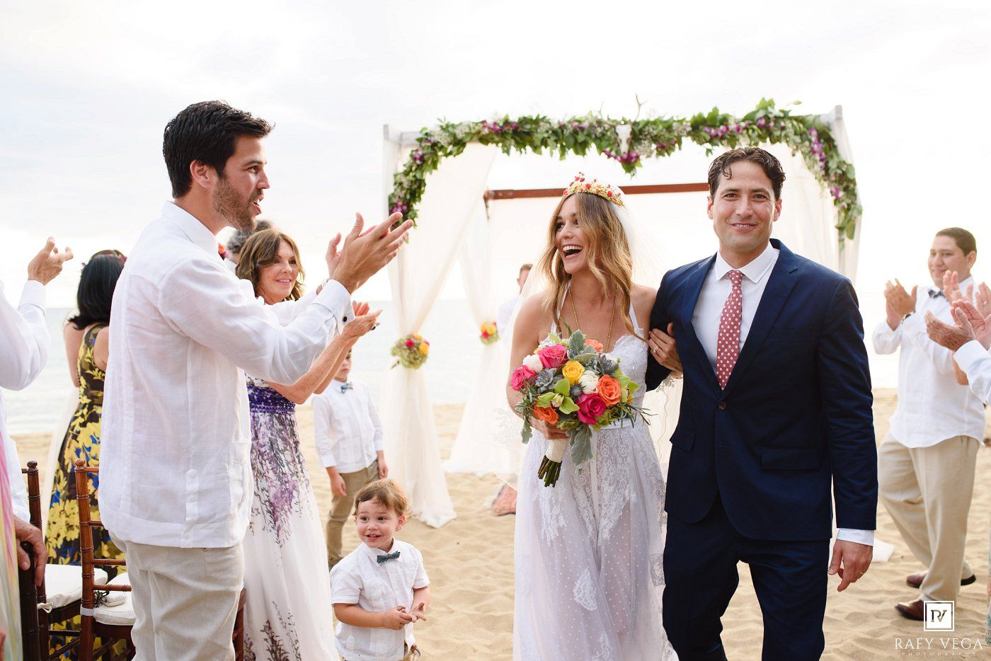 Newlyweds walk down aisle as guests applaud at sunny beach wedding ceremony with floral arch.