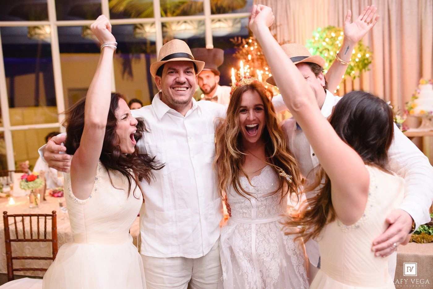 Joyful wedding reception with guests in white attire celebrating with raised arms in a festively decorated venue.