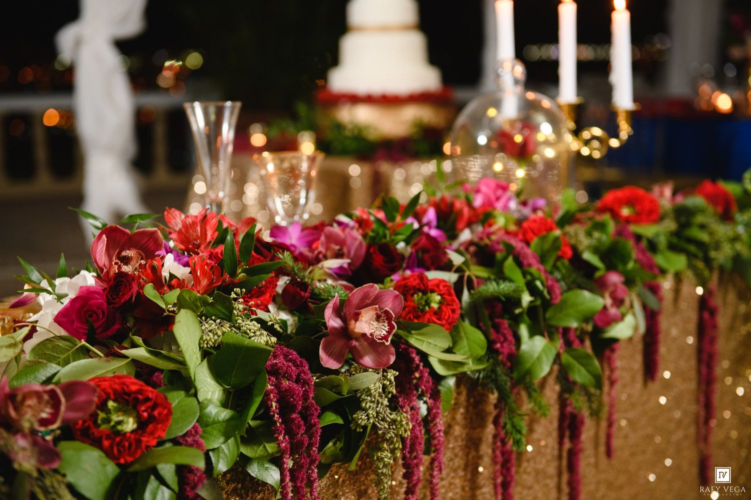 Lush red and burgundy floral centerpiece with greenery on a gold sequin tablecloth at an elegant wedding reception.