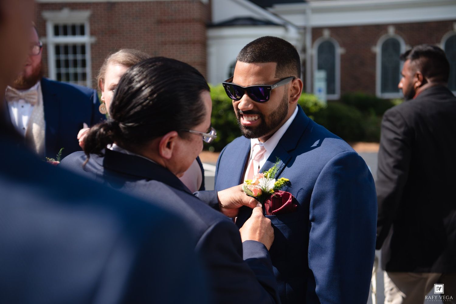 Groom in navy suit and sunglasses gets boutonniere pinned outside a brick church on his wedding day.