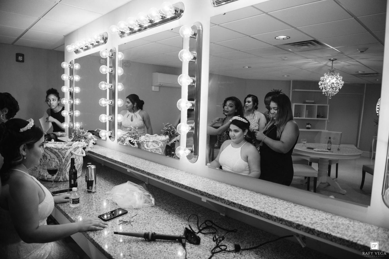 Bridesmaids help a bride get ready in front of a Hollywood vanity mirror in a bridal suite before the wedding.