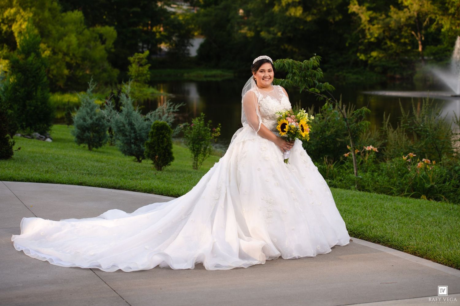 Elegant bride in ball gown with long train holds yellow bouquet outdoors at garden wedding venue.