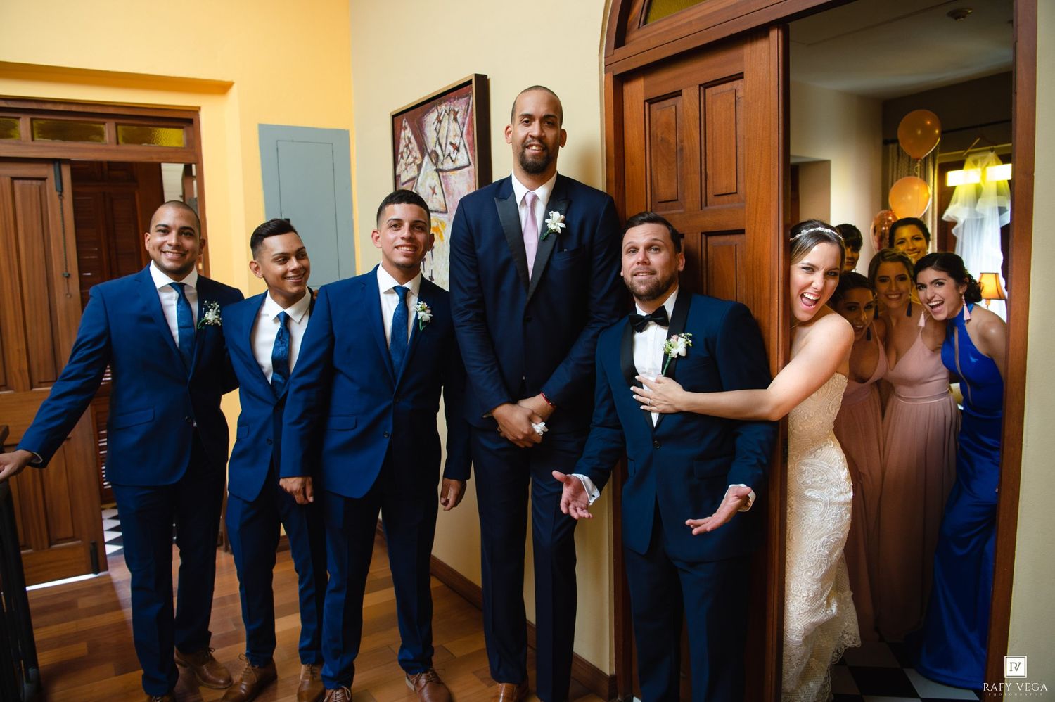 Groom and groomsmen in navy blue suits pose with bride in elegant wedding venue doorway.