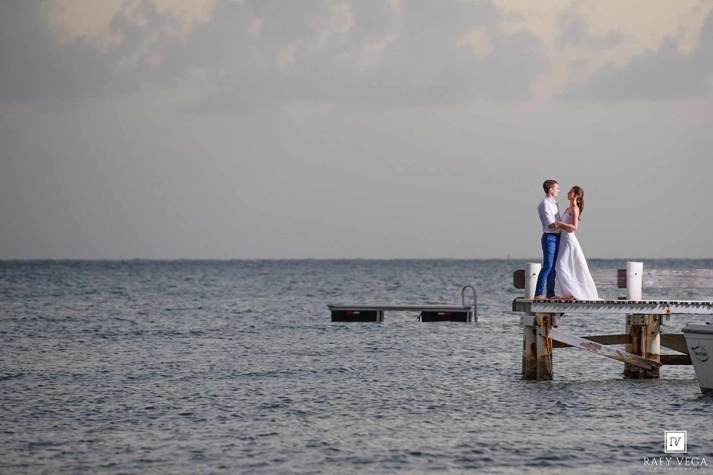 Bride and groom embracing on a wooden pier over calm blue ocean waters under cloudy sky.