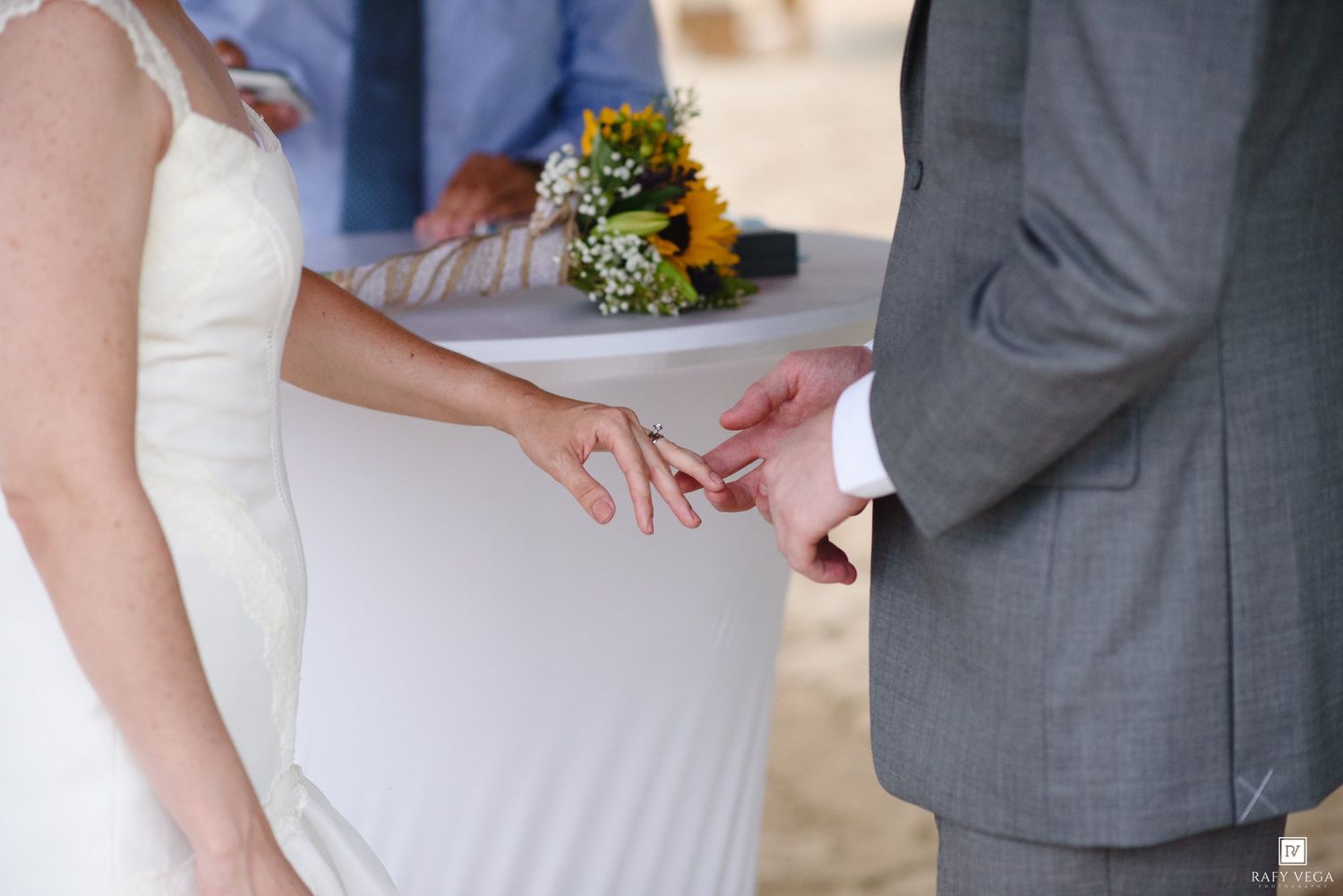 Bride placing a wedding ring on groom's finger during outdoor ceremony, with sunflower bouquet in background.