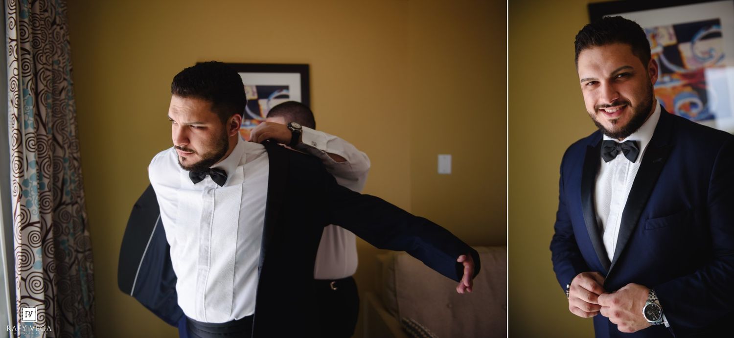 Groom in navy suit and bow tie being helped by groomsman while smiling during wedding day preparations.