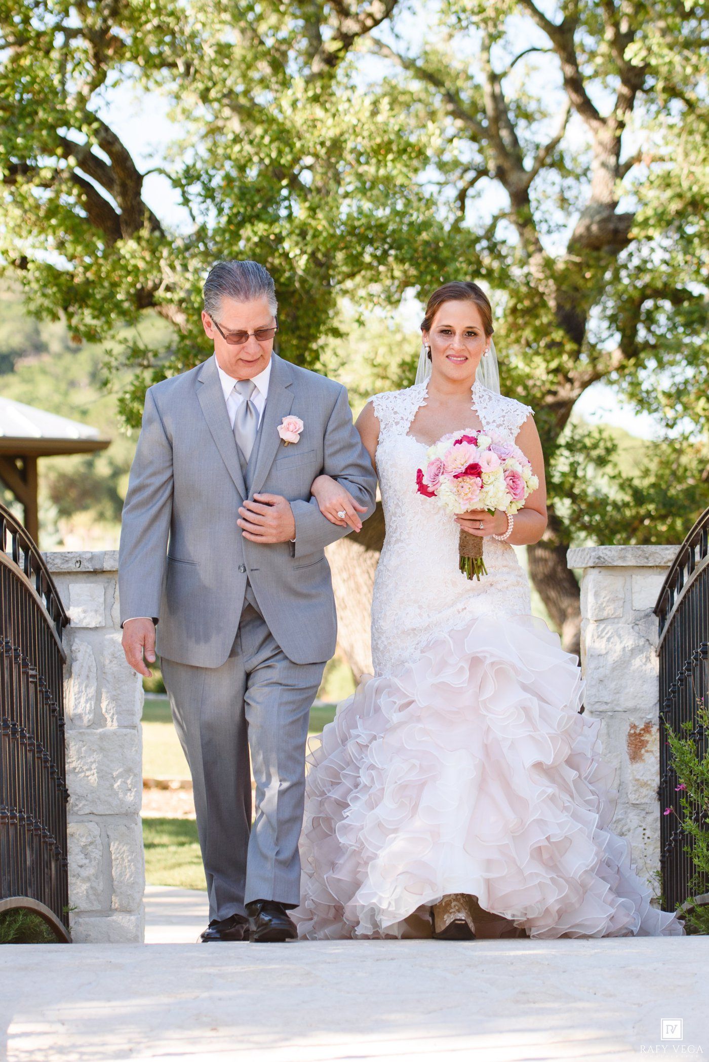 Bride in ruffled gown walks with father in gray suit on outdoor bridge, holding pink bouquet at wedding ceremony.
