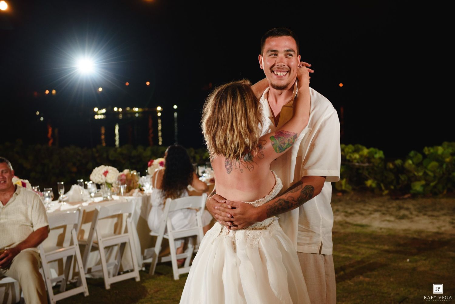 Groom in white linen shirt lifts bride during outdoor beach wedding reception at night.