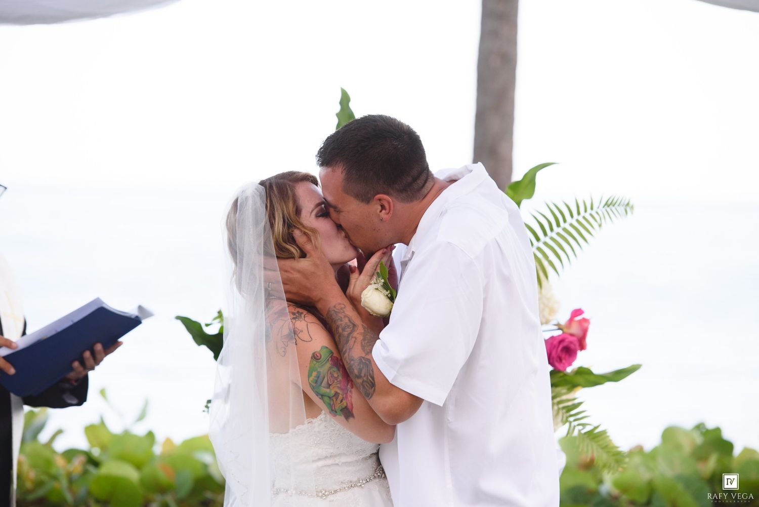 Bride and groom share their first kiss at an outdoor beach wedding ceremony with tropical flowers and ocean views.