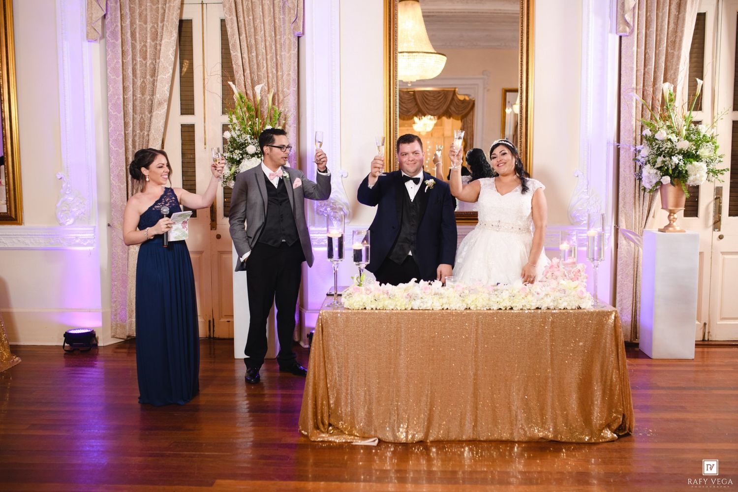 Wedding reception toast with bride, groom, and bridesmaid raising glasses at gold-draped head table in elegant ballroom.