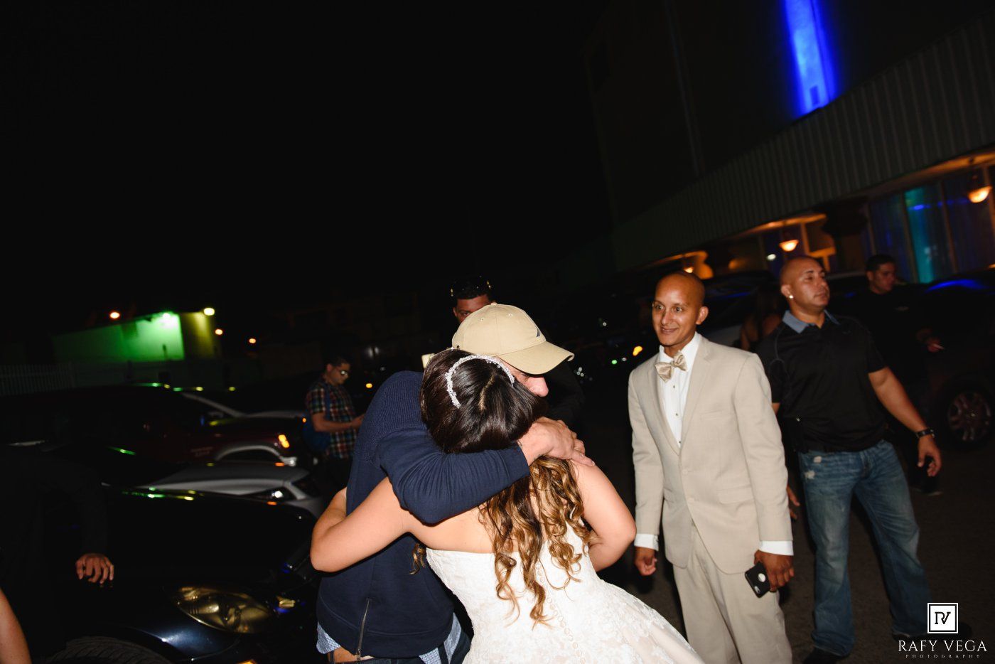 Bride in white gown receives a warm hug outdoors at night during a wedding celebration.