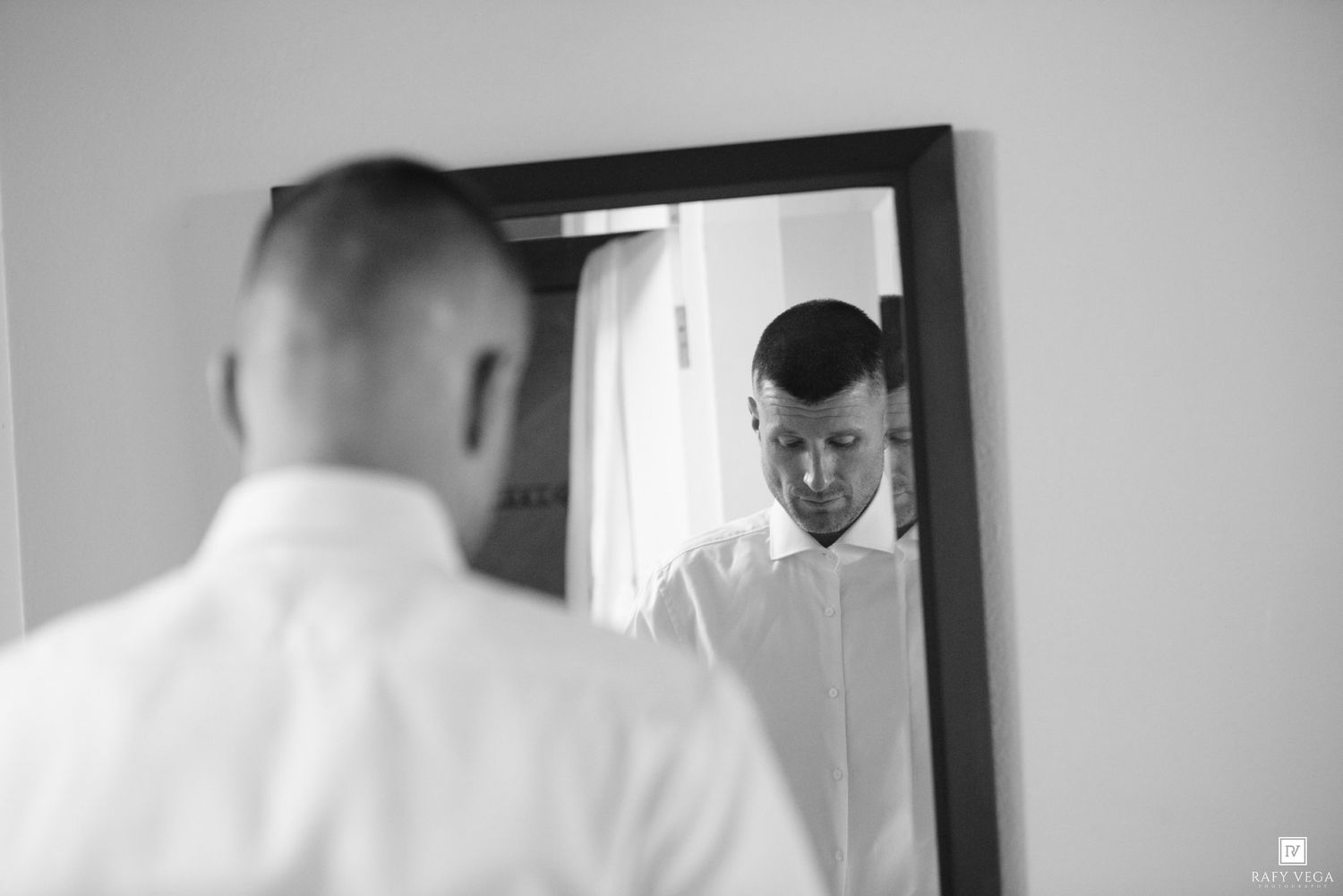 Groom in white dress shirt adjusting his outfit while looking down, reflected in a mirror in black and white.