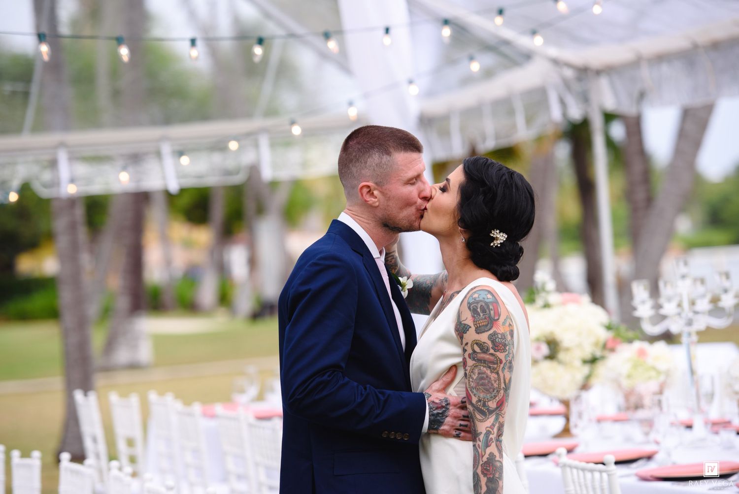 Bride and groom share a passionate kiss at their outdoor wedding reception under a festive tent.