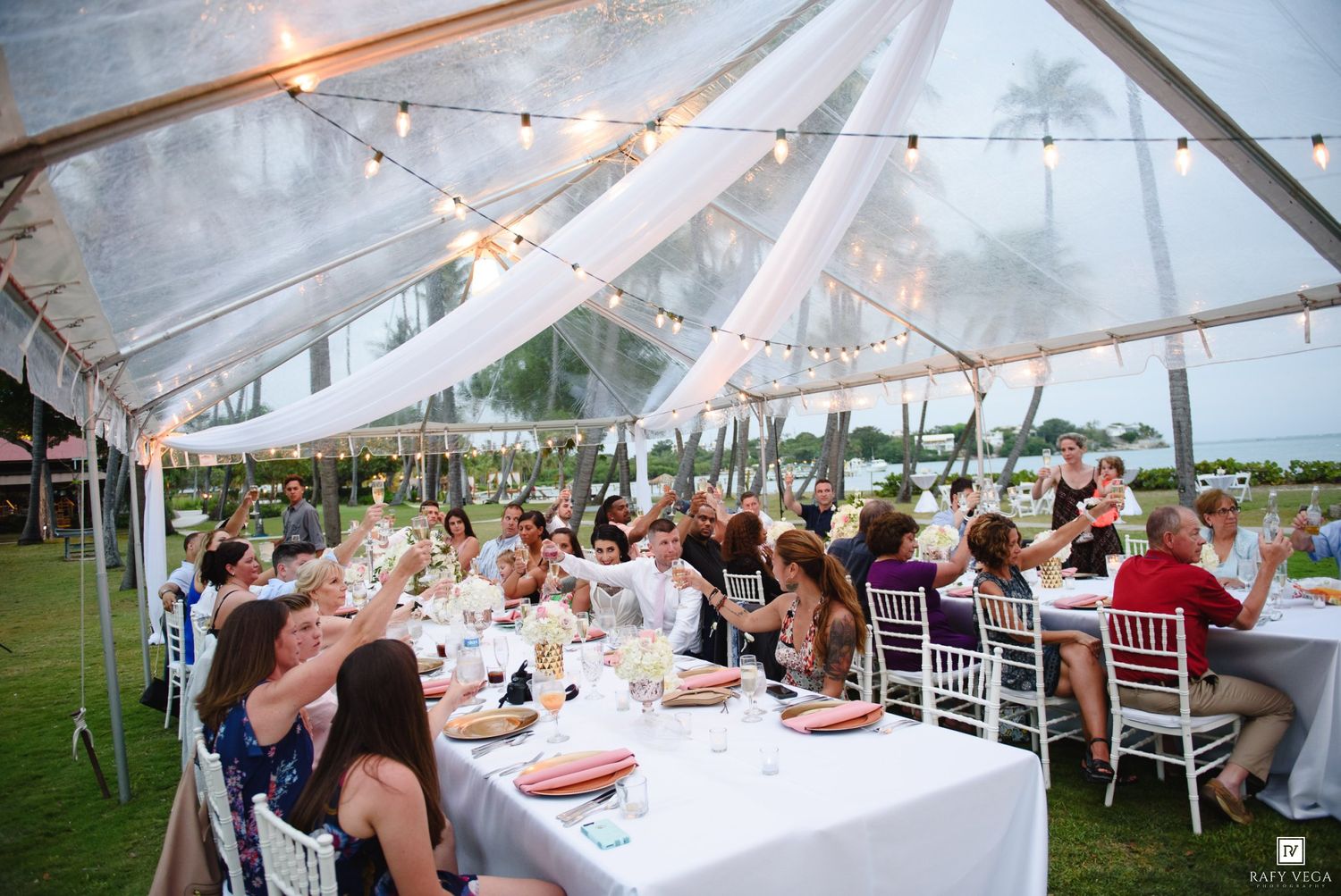 Guests dining under a white tent with string lights at an outdoor wedding reception on a tropical lawn.