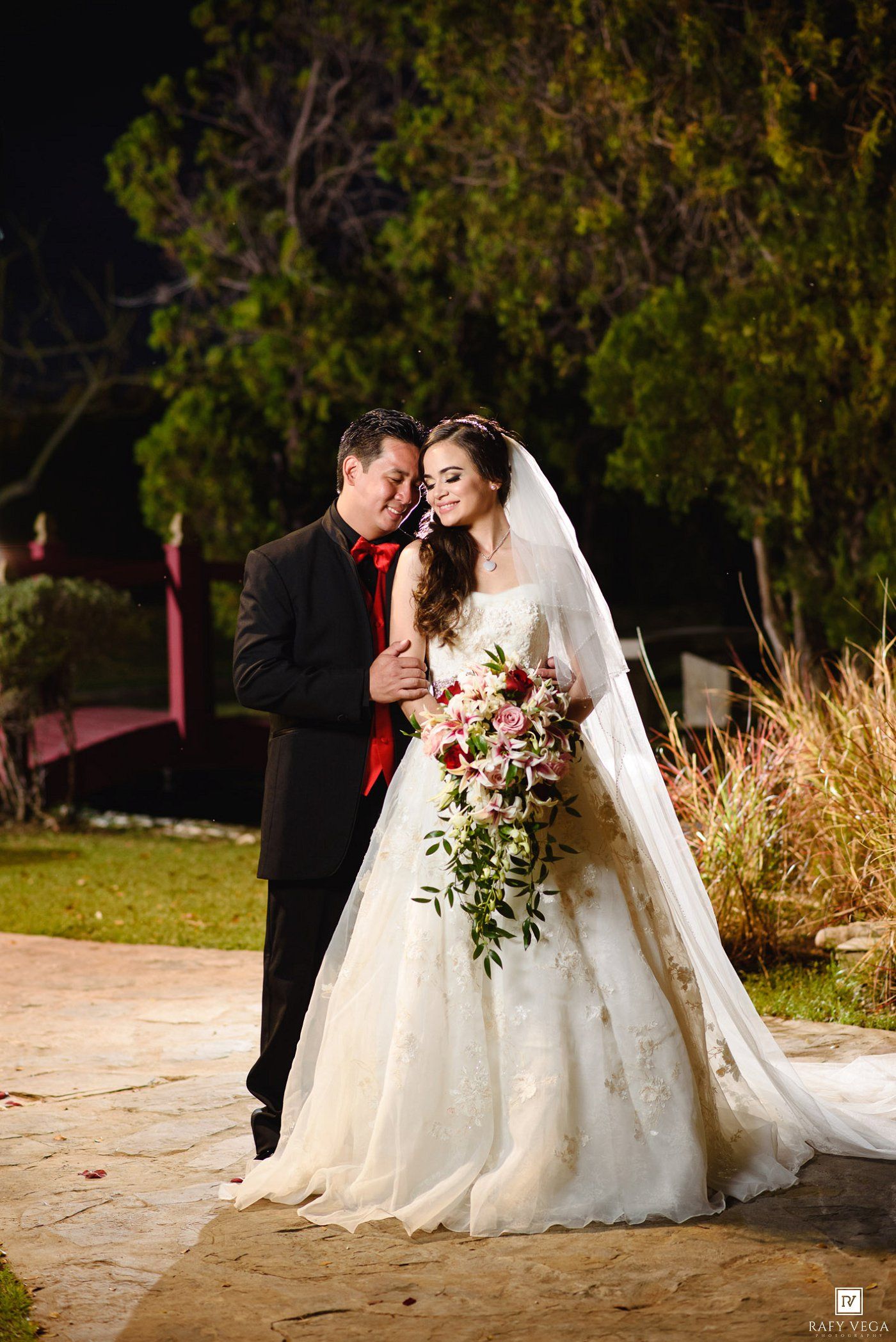 Bride in white gown with bouquet and groom in black tuxedo pose together outdoors at night wedding celebration.